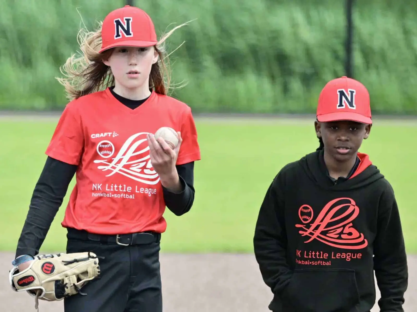 Twee jonge honkballers in rode NK Little League-caps lopen het veld af, een met bal en handschoen.