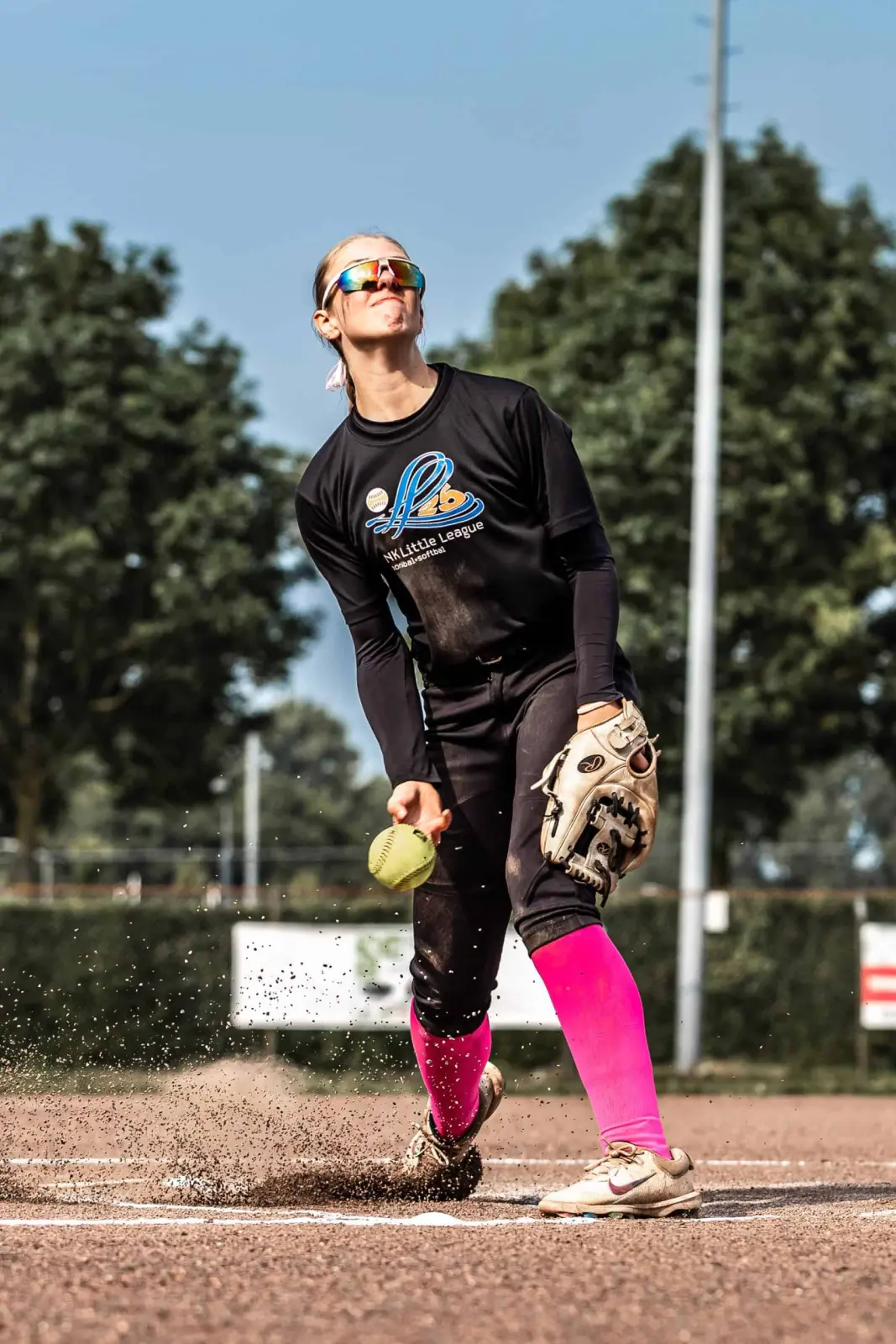 Softbalpitcher in zwart shirt en roze kousen werpt onderhands een gele bal, het gravel stuift op.