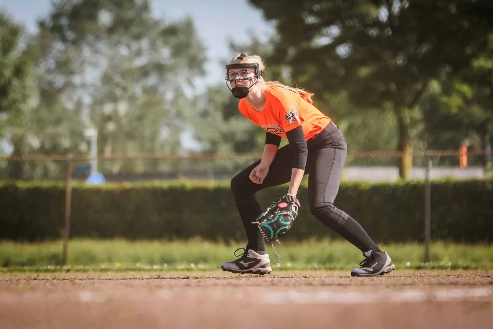 Softbalspeelster in oranje shirt en gezichtsmasker staat gebukt klaar met haar handschoen op het binnenveld.