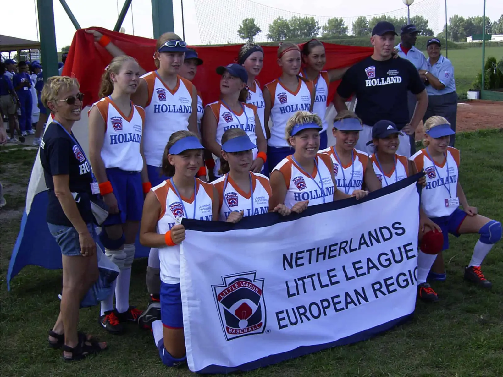 Softbalteam in Holland-shirts poseert lachend met een Nederlandse vlag en een banner van Little League European Region.