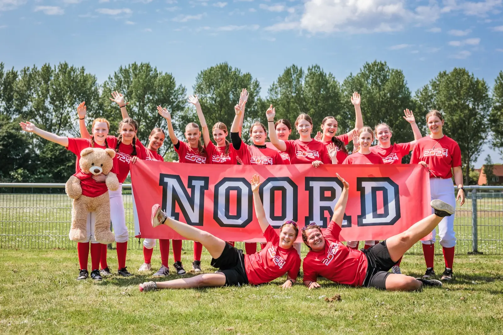 Het softbalteam van regio Noord juicht achter een spandoek op het veld tijdens het NK Little League 2024.