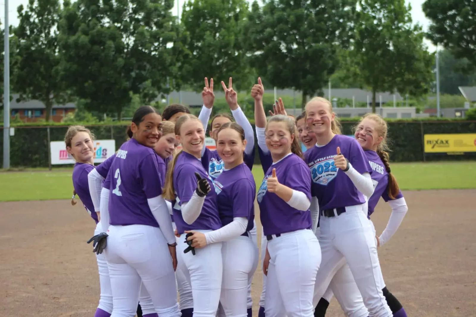 Softbalteam in paarse Vipers-shirts juicht met duimen omhoog en vredestekens op een gravelveld.
