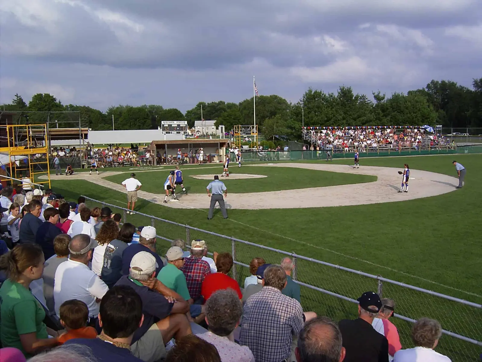 Softbalwedstrijd gezien vanaf een volle tribune, met het binnenveld, umpires en een scorebord op de achtergrond.