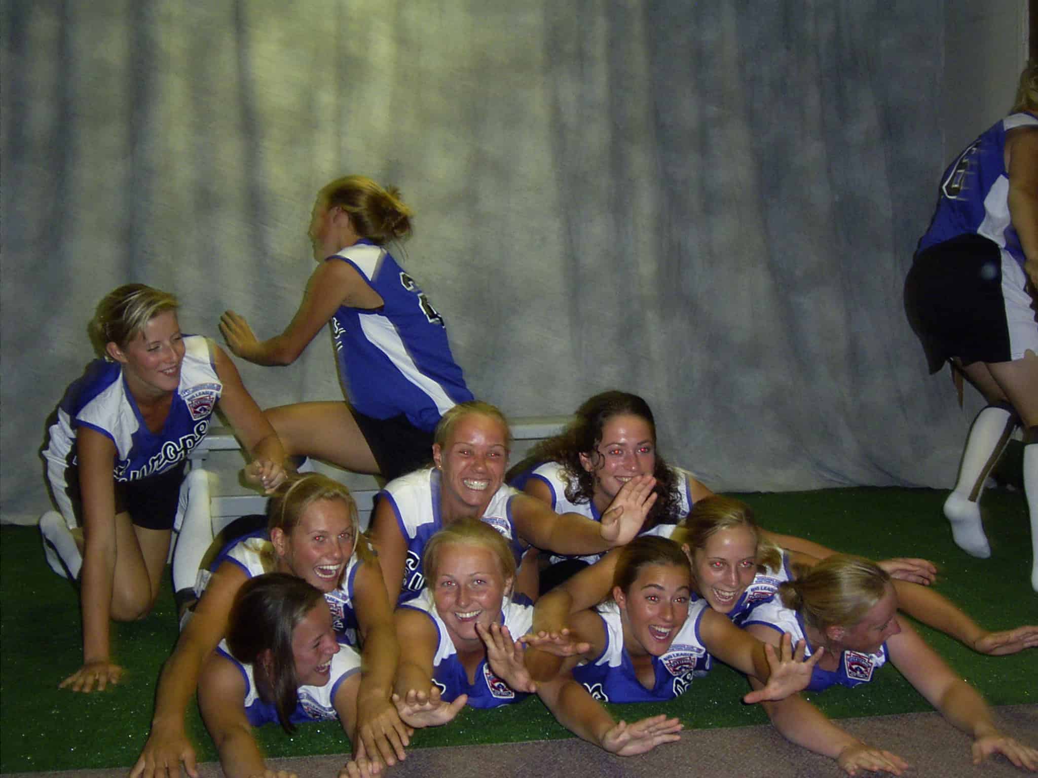 Softbalspeelsters van Team Europe in blauw-witte tenues liggen lachend op een stapel op het kunstgras.