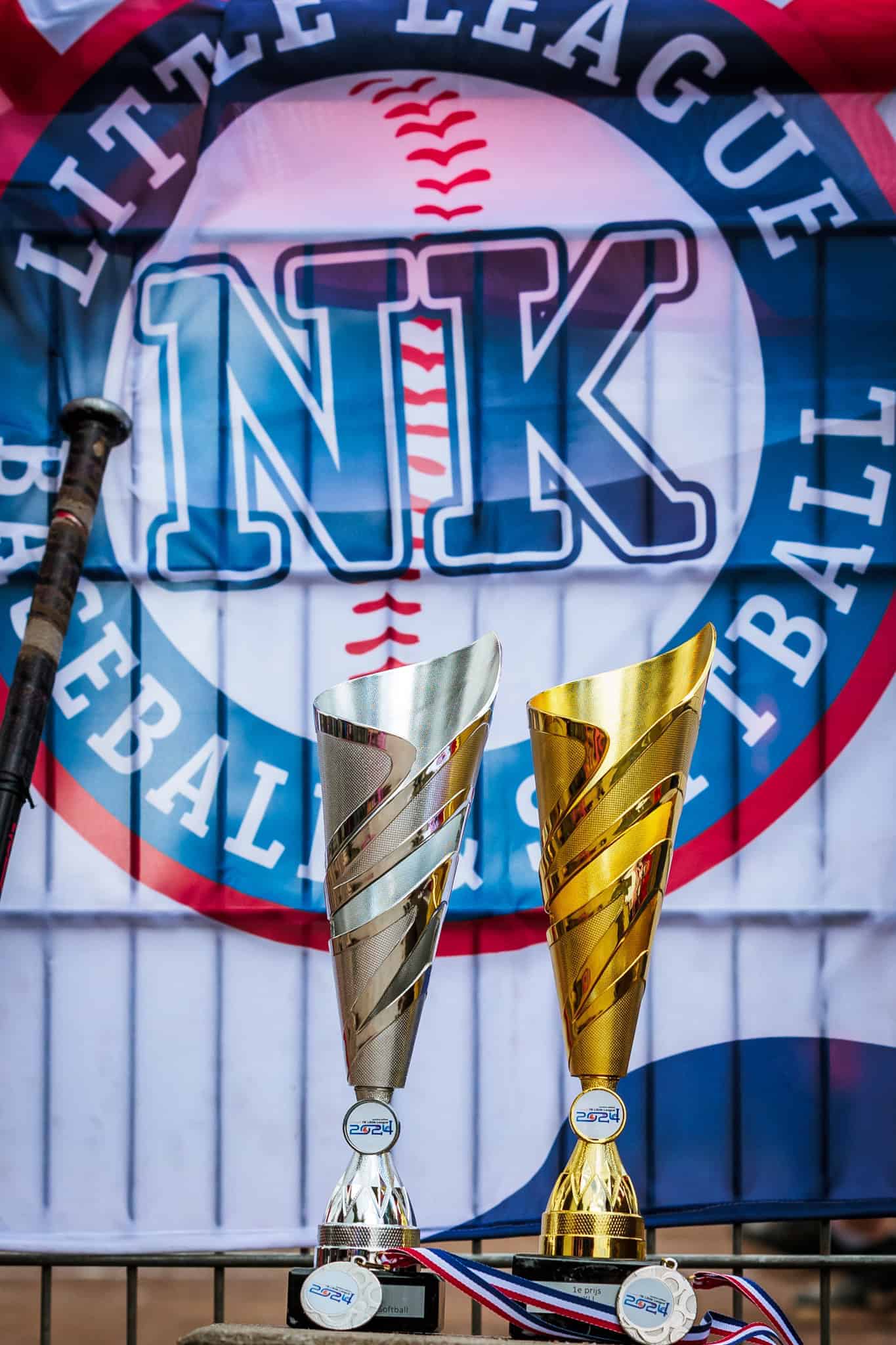 Een zilveren en een gouden wisselbeker met medailles voor de vlag van het NK Little League.