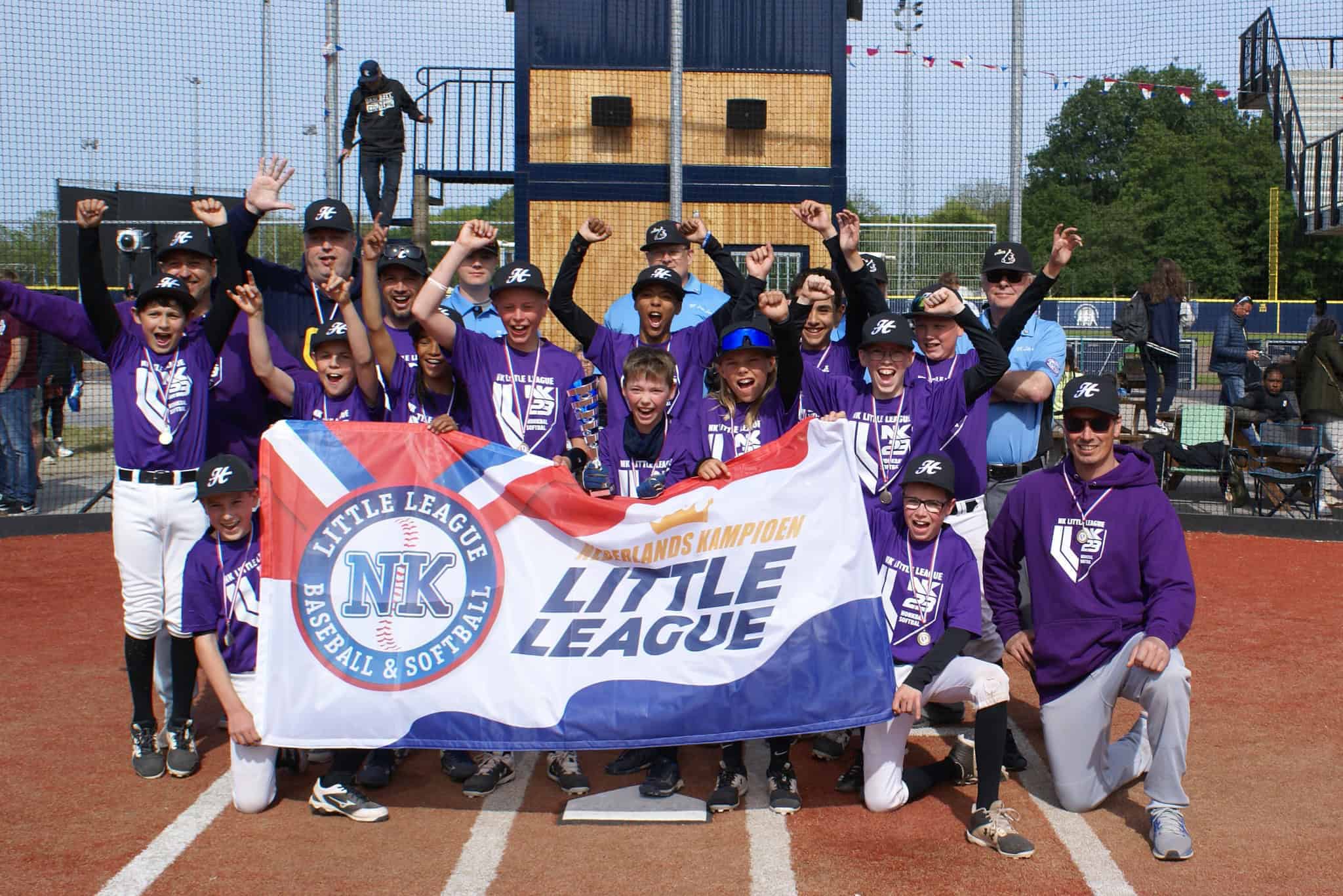 Juichend jeugdhonkbalteam in paarse shirts met de vlag Nederlands Kampioen Little League op het veld.