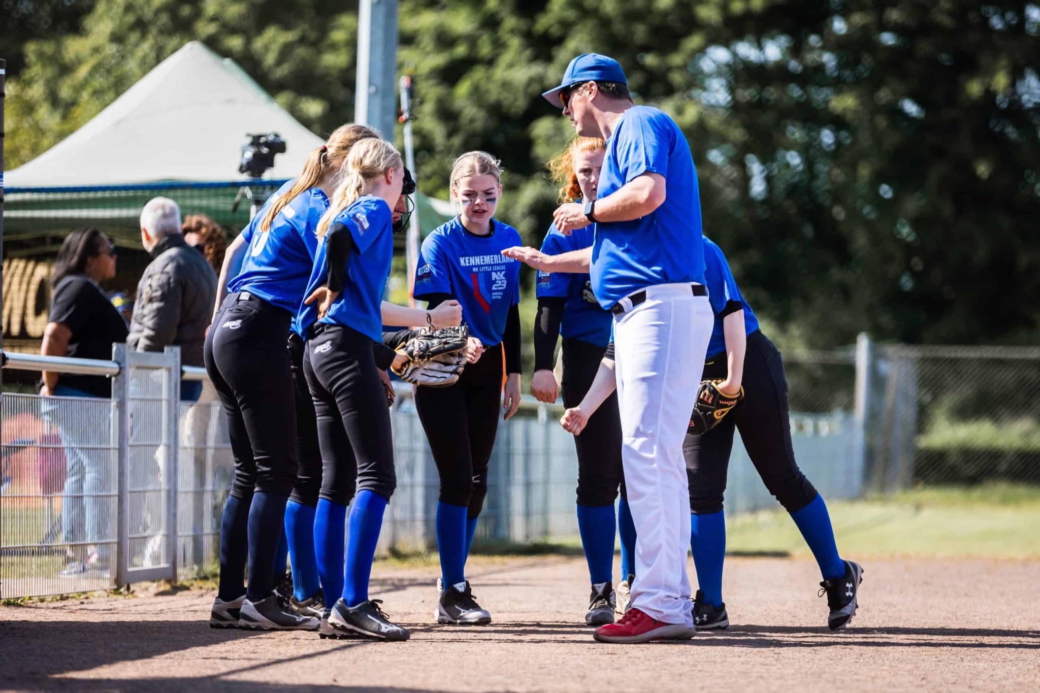 Coach in blauw shirt spreekt een groep softbalspeelsters van Kennemerland toe naast het veld.