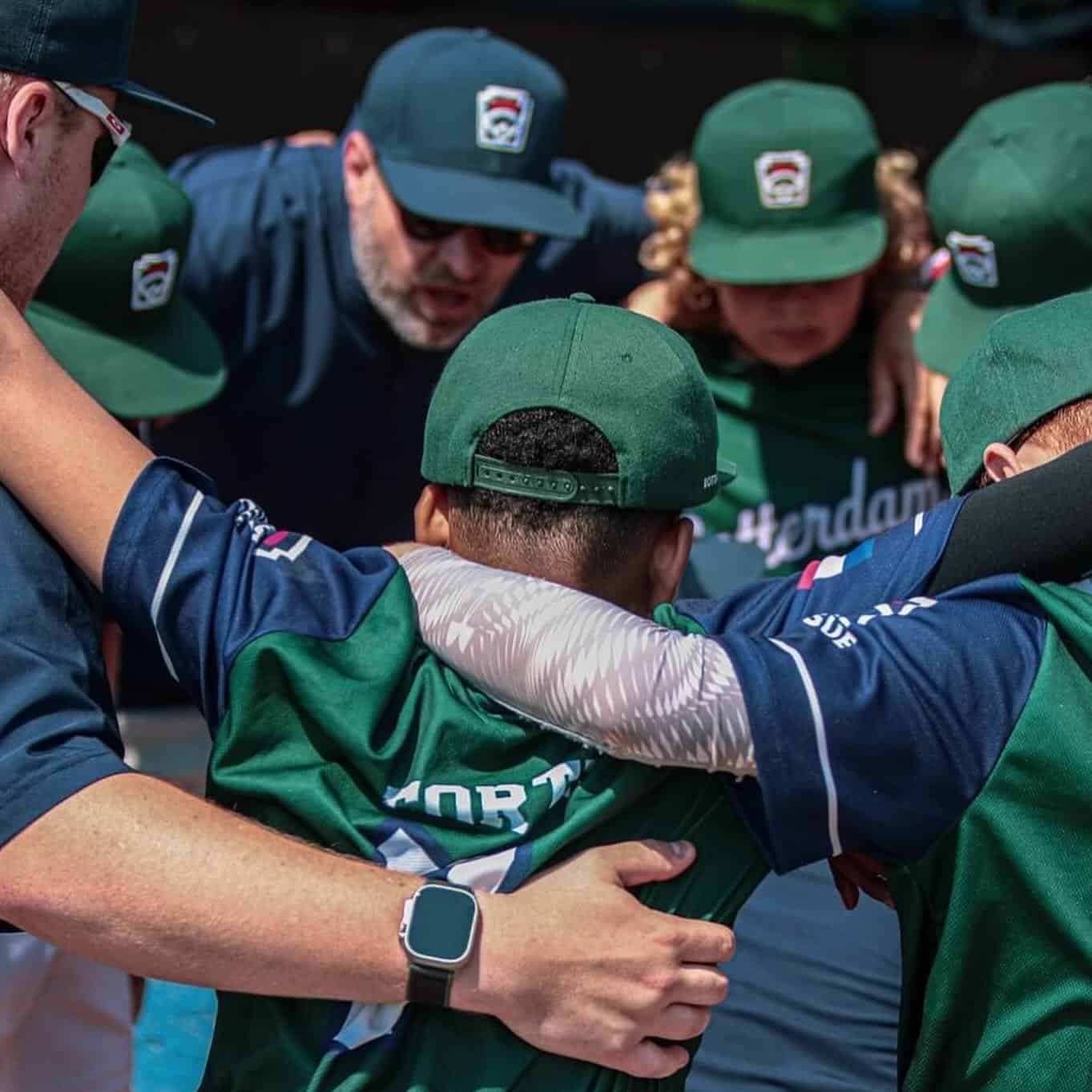 Spelers en coaches van Rotterdam met groene petten staan in een kring met de armen om elkaar heen.