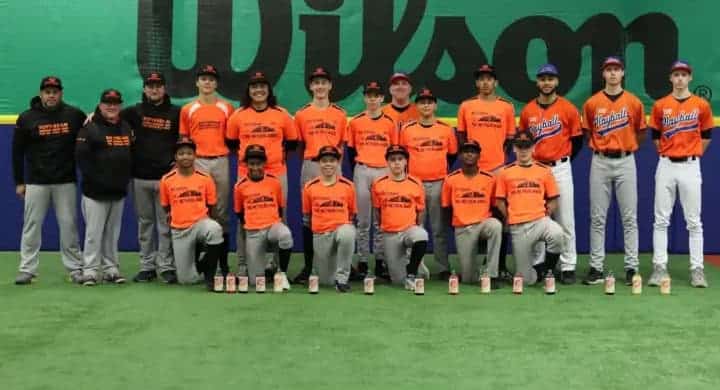Teamfoto van een honkbalteam in oranje shirts met coaches in een indoorhal.