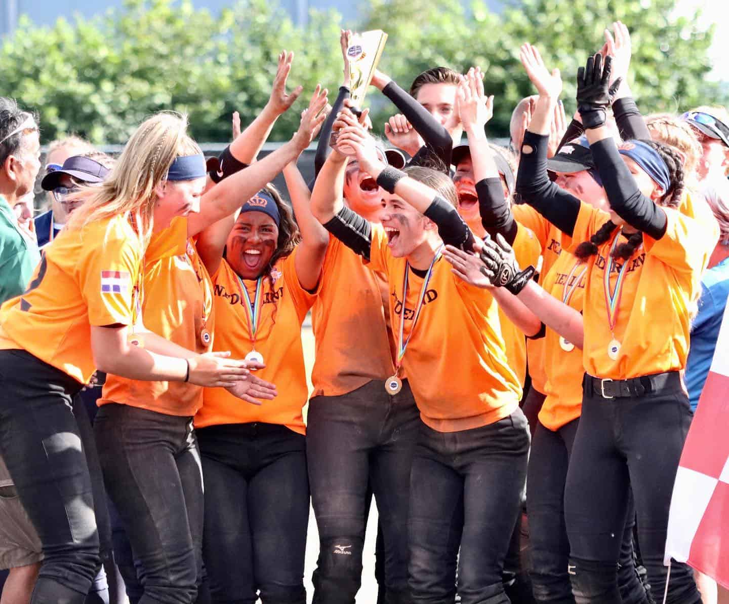 Softbalsters in oranje shirts met medailles om juichen met de armen omhoog rond de beker.