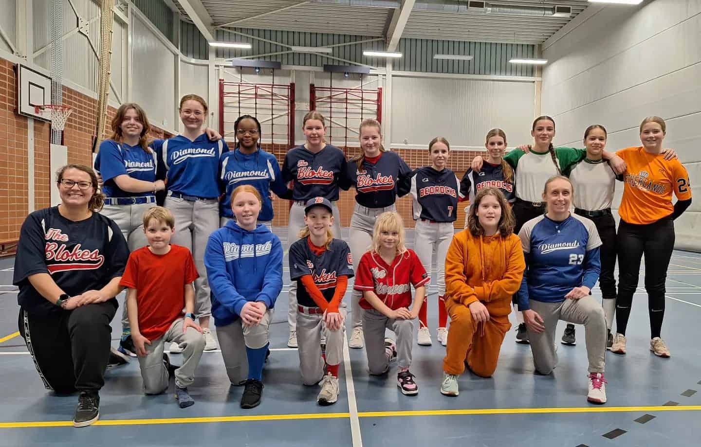Groepsfoto van softbalspeelsters van verschillende verenigingen in een sporthal.