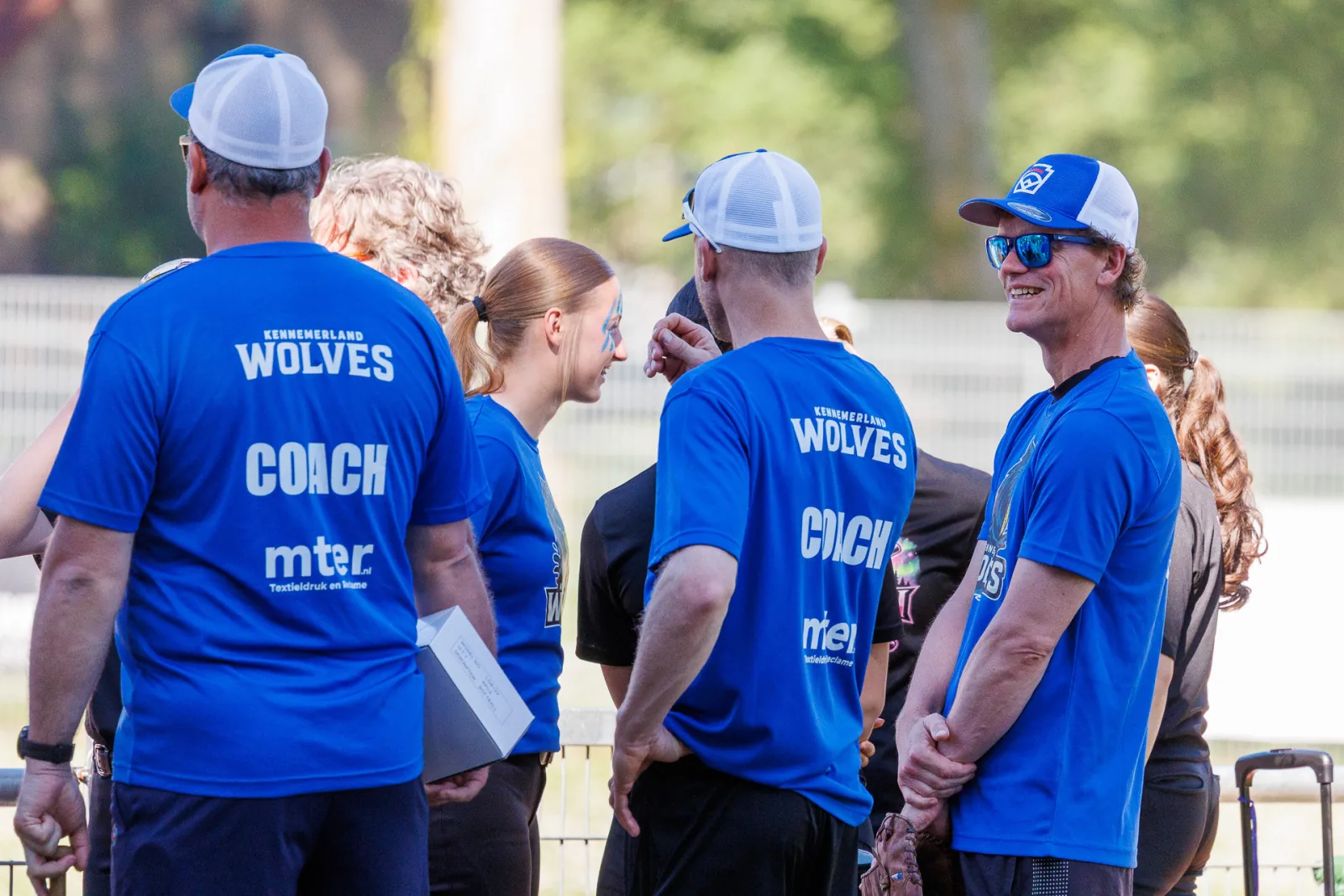 Coaches in blauwe coachshirts overleggen langs het veld.