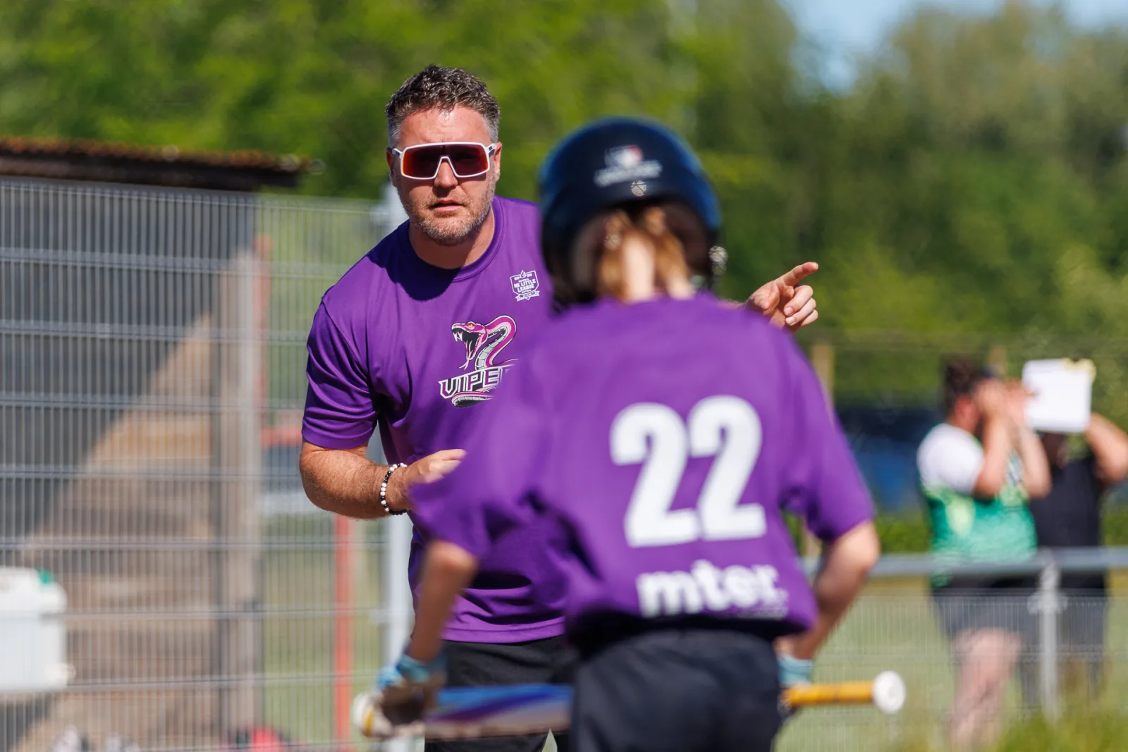 Een coach in een paars shirt geeft aanwijzingen aan een slagman met helm.