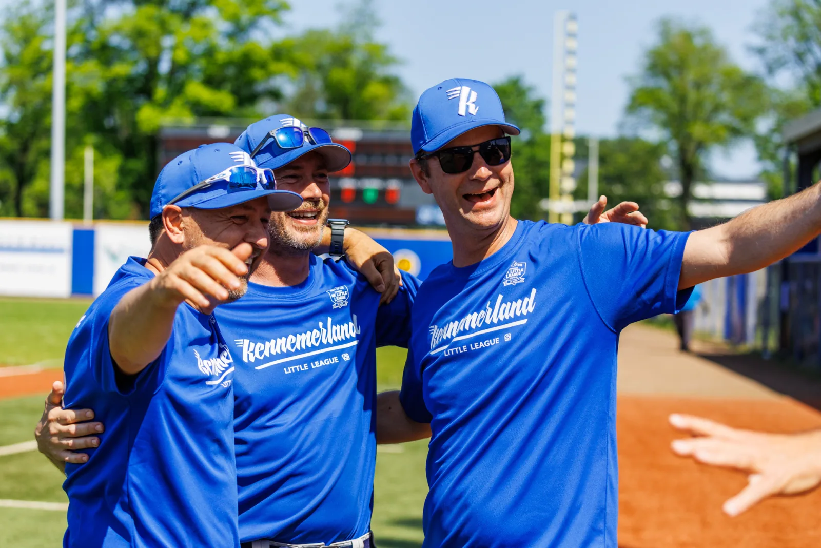 Drie coaches van Kennemerland in blauwe shirts staan lachend met de armen om elkaar heen langs het veld.