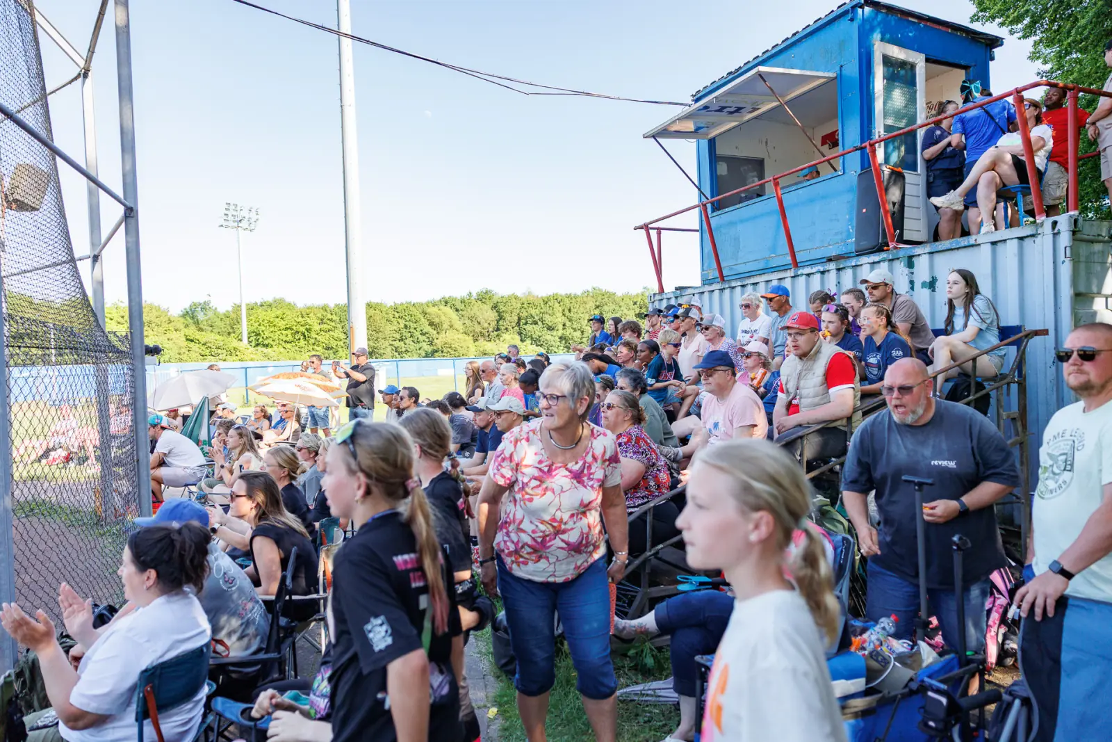 Volle tribune langs het veld; supporters kijken naar de wedstrijd vanaf de banken bij het scorebordhuis.