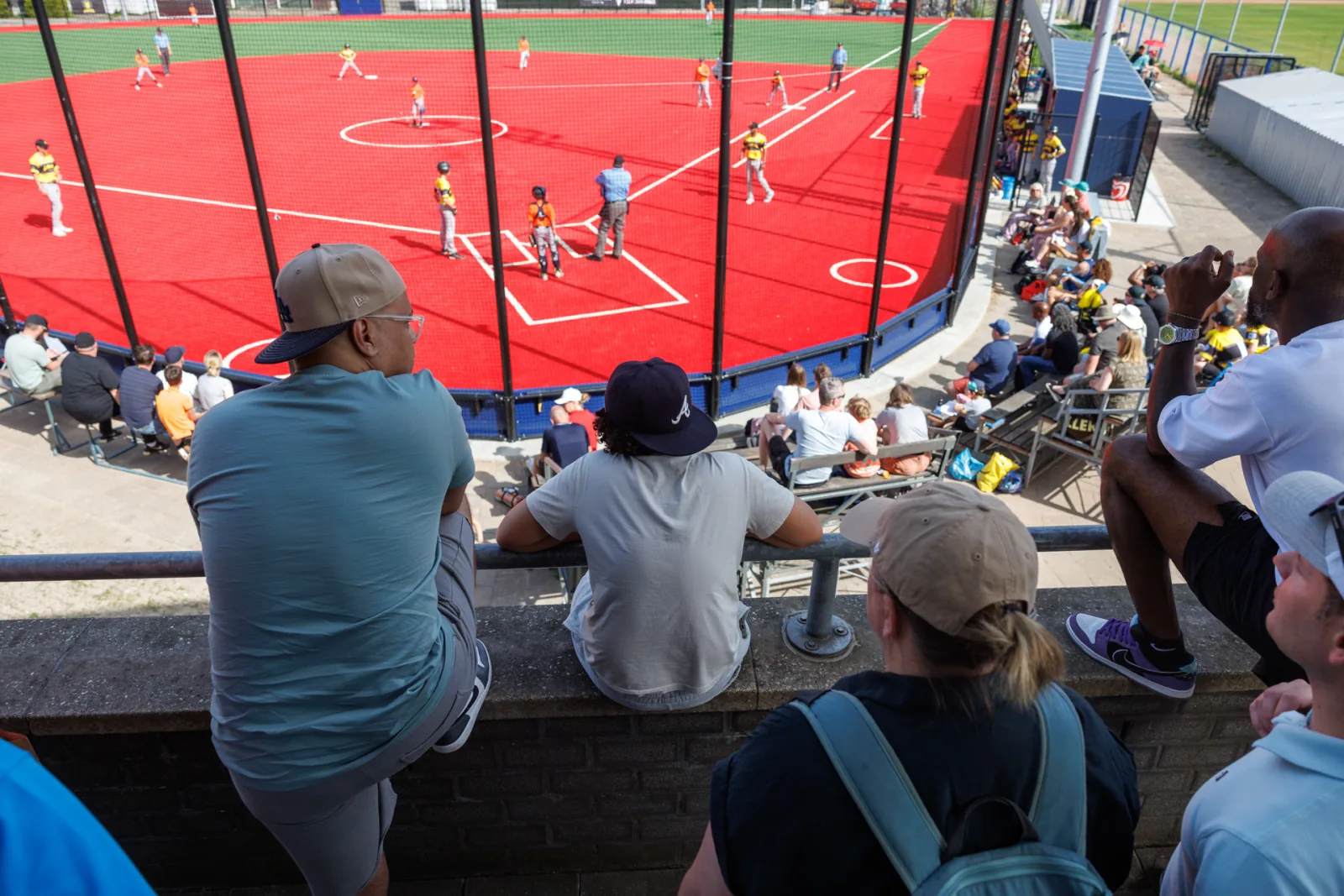 Publiek kijkt vanaf de tribune uit over het rode softbalveld.