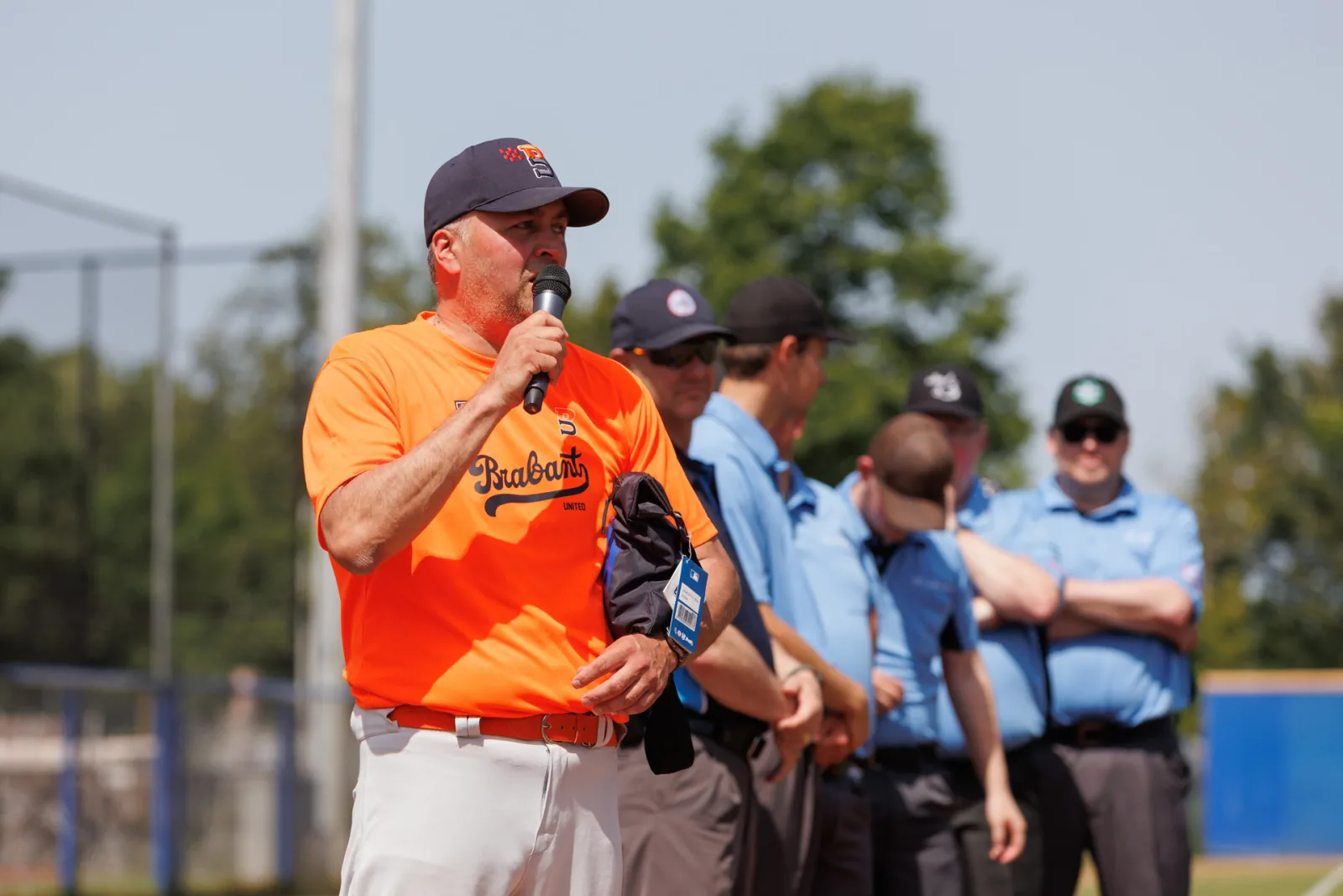 Iemand van de toernooileiding spreekt het veld toe met een microfoon, umpires luisteren mee.