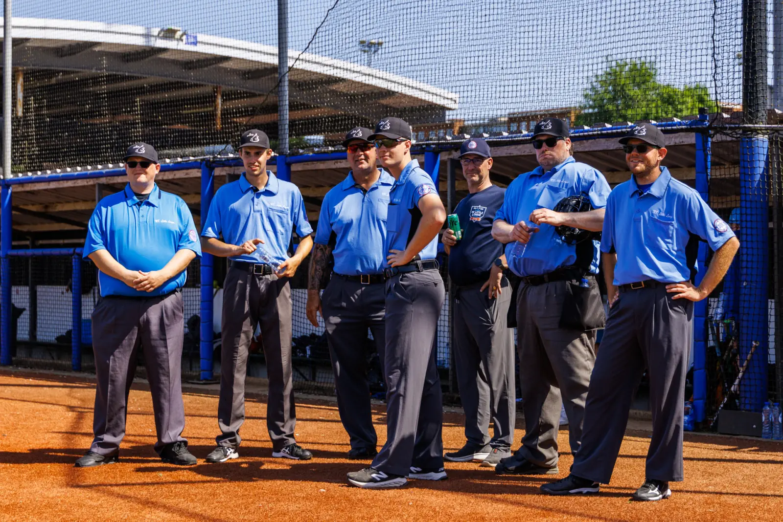 Zeven umpires in lichtblauwe shirts staan bij elkaar op het gravel voor de dug-out.